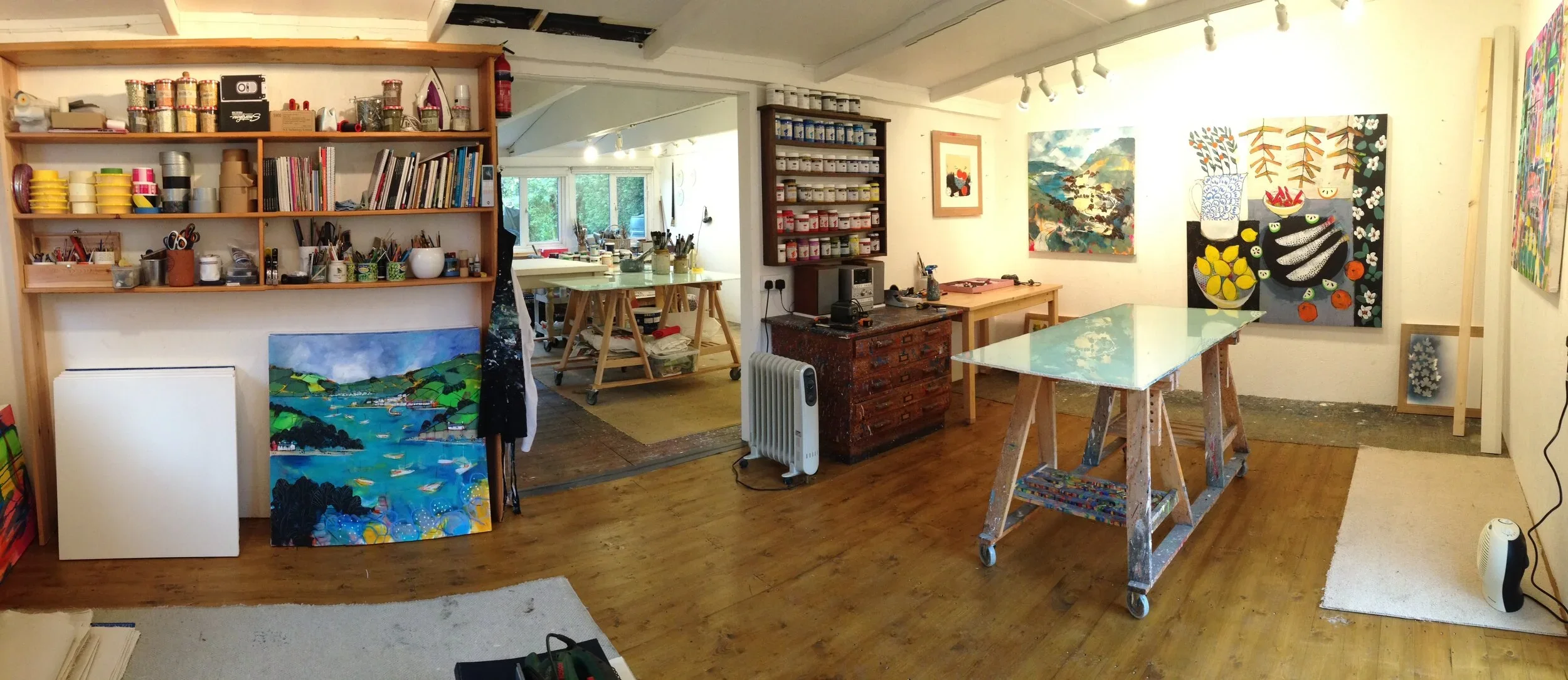 An art studio with paintings on the walls, a large wooden table covered in paint splatters, shelves with art supplies, and a workspace with a pottery wheel. Natural light streams in from windows.
