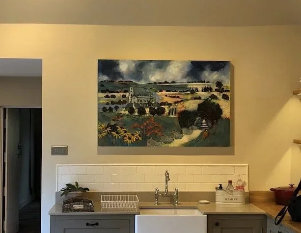 Painting of a rural landscape with fields, trees, and a cloudy sky hung above a kitchen sink area.
