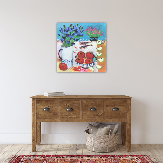 Colorful still life painting of flowers, fruits, and a pitcher hanging on a plain white wall above a wooden console table with books and a woven basket.