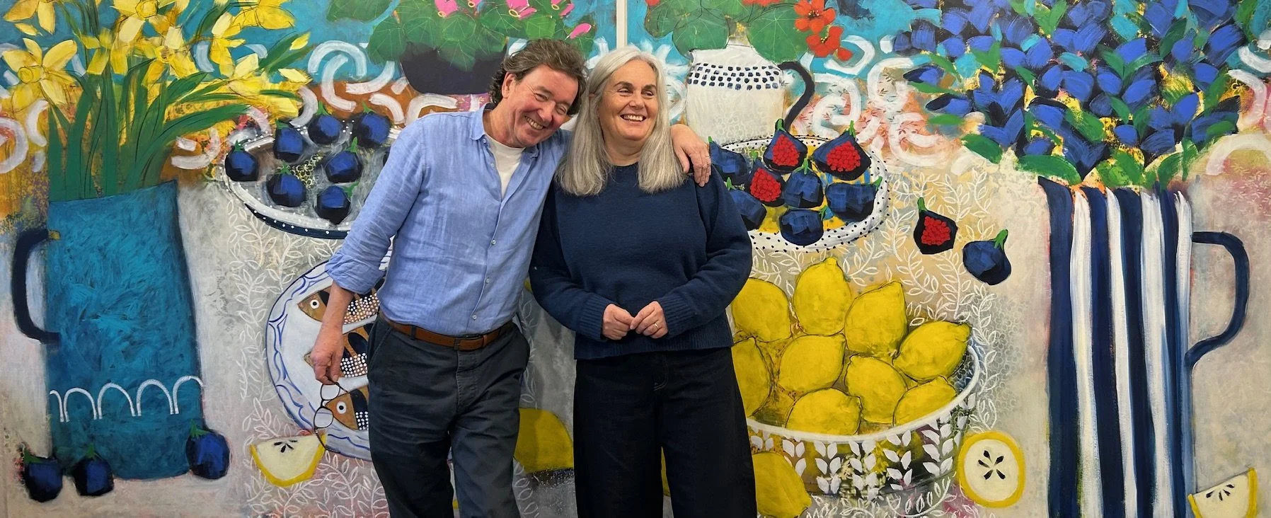 Two people smiling and standing close together in front of a colorful, painted mural of various household items and flowers.
