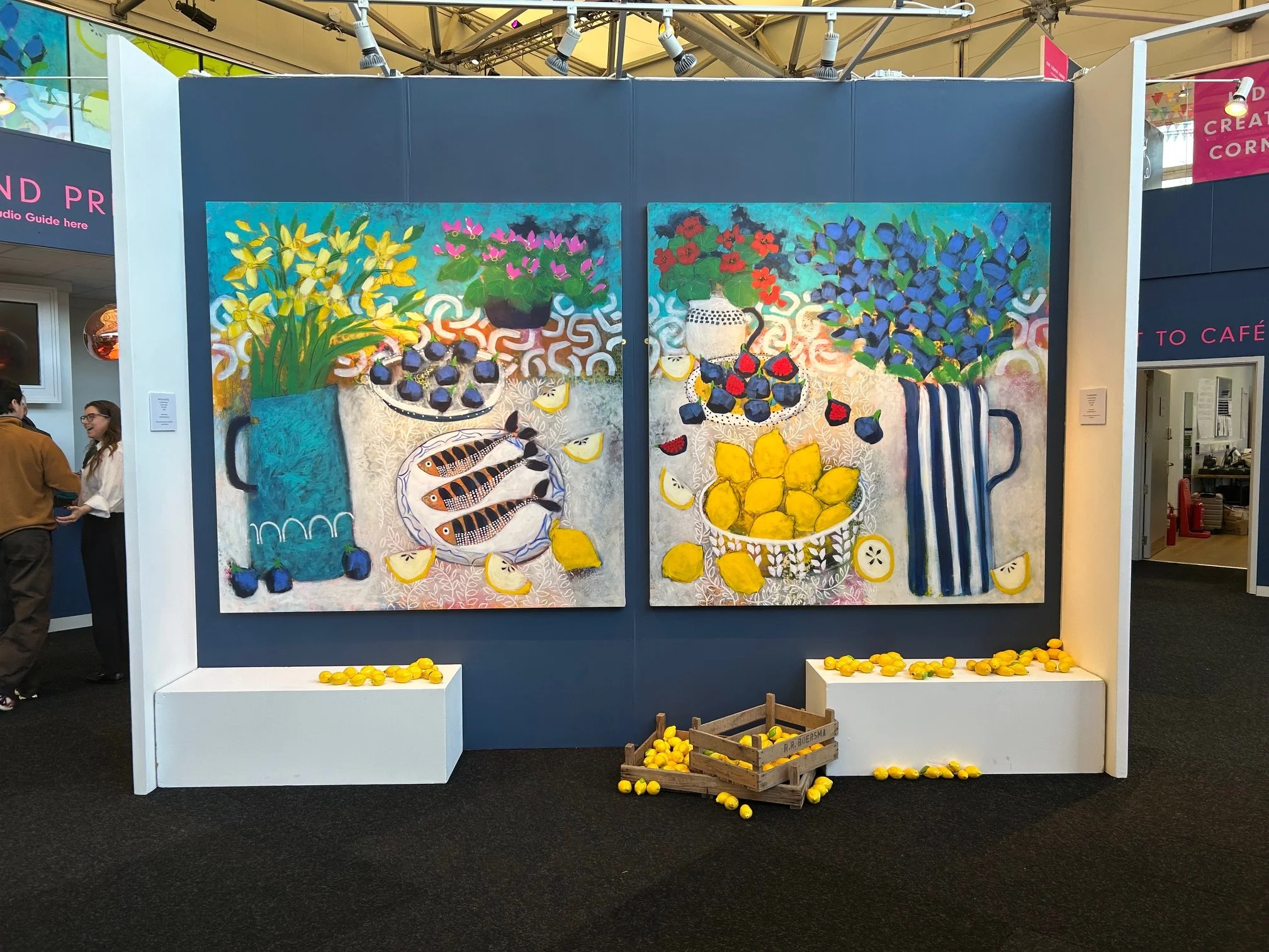 An indoor art display features two colorful paintings of still life flowers and fruit arranged on the wall, with small yellow lemons arranged in front and around the base of the display.