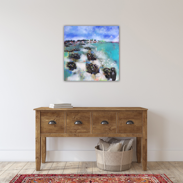 Painting of a seaside landscape with a blue sky, water, and rocks, hanging above a wooden console table with drawers, decorated with a stack of books and a woven basket on the floor.