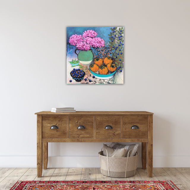Colorful still life painting of flowers and fruit hanging on a white wall above a wooden console table, with a basket of blankets on the floor.