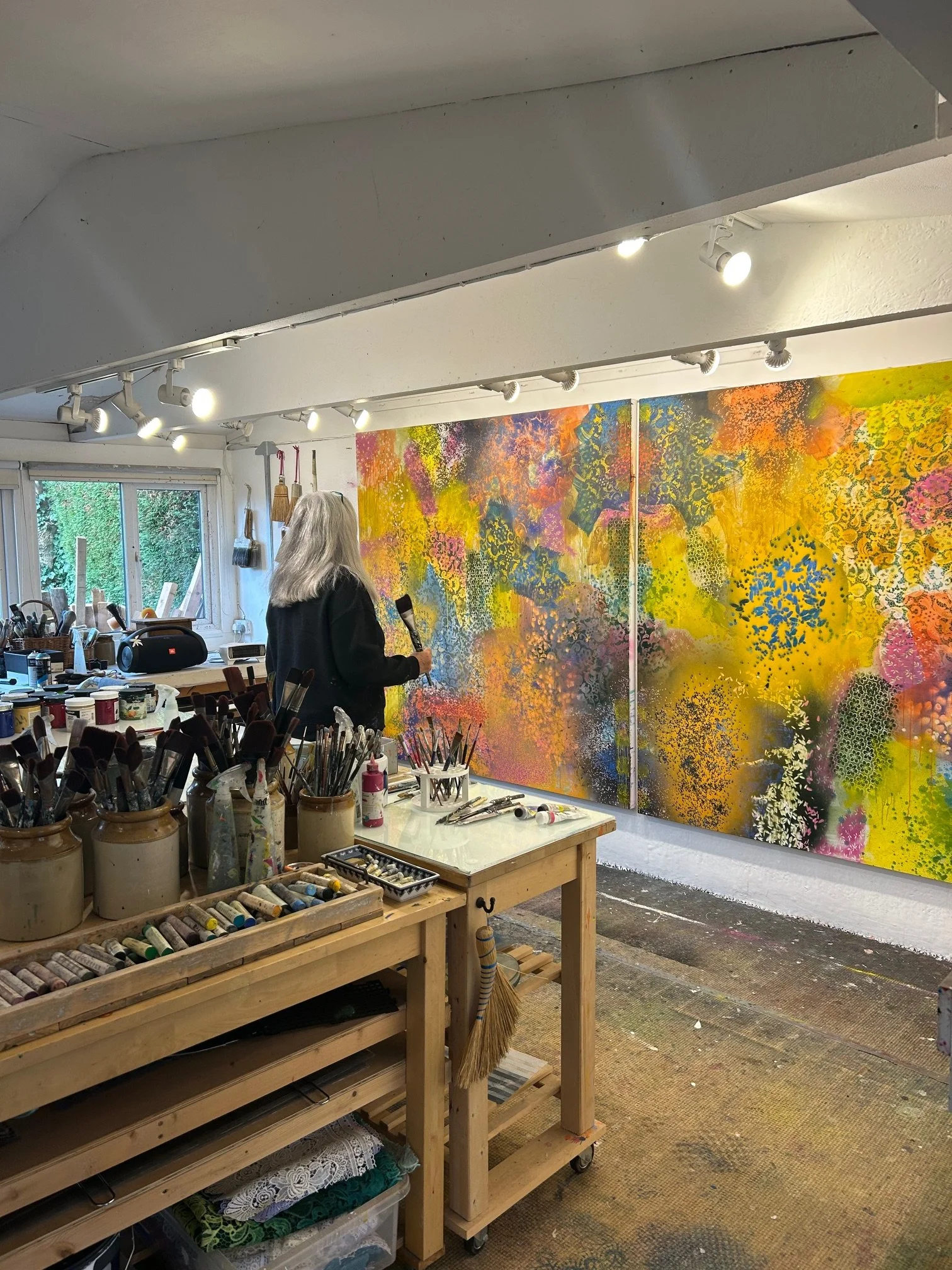 An artist creating a colorful abstract mural on a wall, with painting supplies and brushes on a nearby table, and a woman holding a microphone observing the artwork.