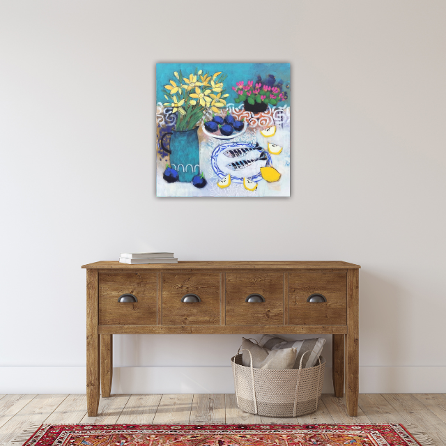 Colorful still life painting of flowers and a fish on a white wall above a wooden console table with books and a storage basket underneath.