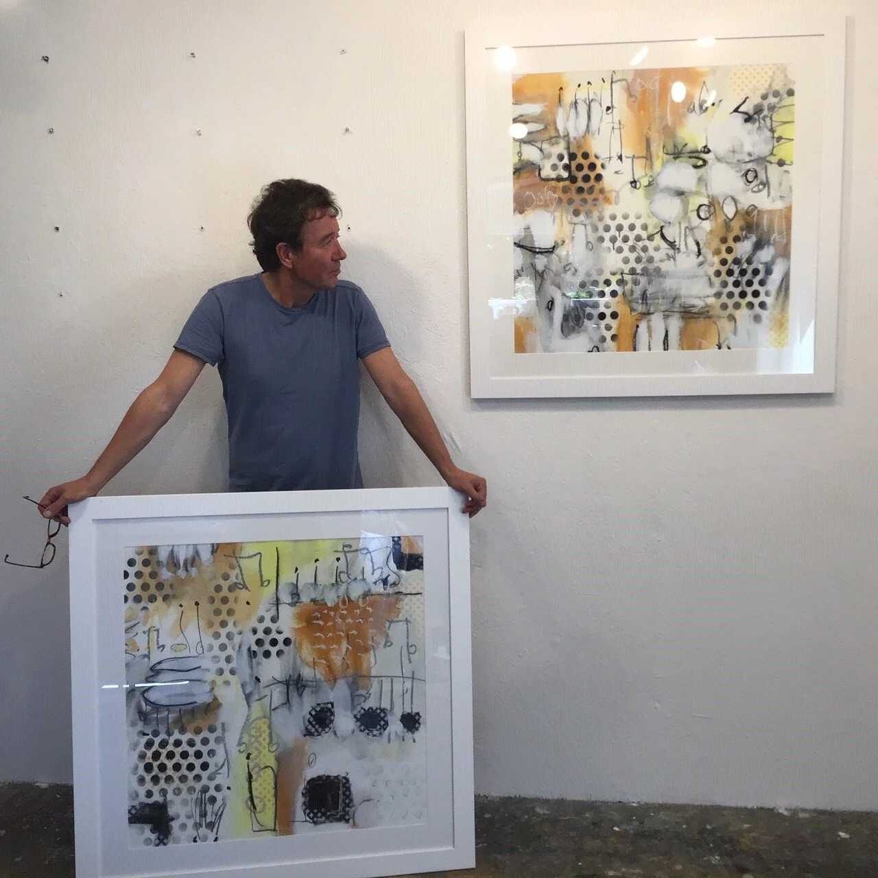 A man holding a framed abstract artwork in front of him with a similar artwork hanging on the wall behind.