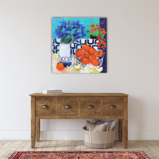 Colorful still life painting of vases with blue, red, green, and orange flowers on a table, hanging on a white wall above a wooden console table with three drawers, a basket with blankets, and a red patterned rug in a room.
