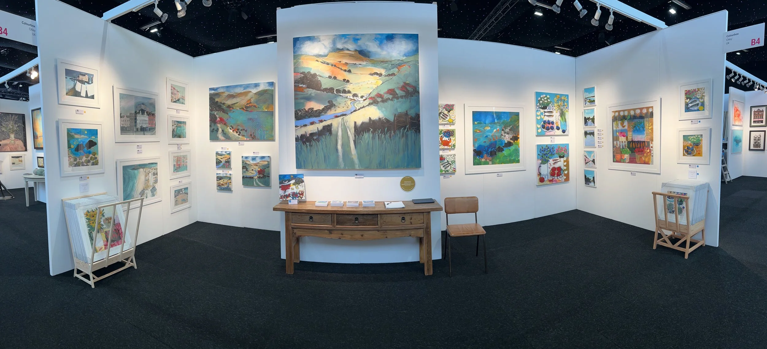Art gallery with white walls displaying colorful paintings, including landscapes and abstract artwork. There are small tables and chairs, with art catalogs on the tables and a ladder in the corner.