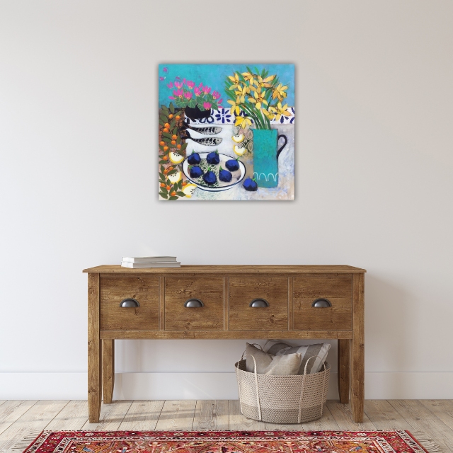 Colorful still life painting of fruit and flowers hung on a white wall, above a wooden console table with books and a basket on the floor.