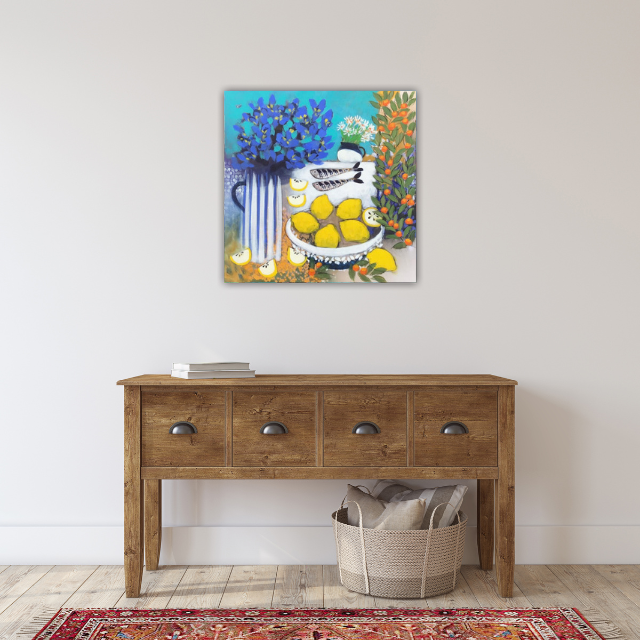 Painting of a blue vase with yellow flowers, a white teapot, and lemons on a table, hung on a plain white wall above a wooden console table with four drawers and a basket of pillows underneath.