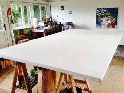 A large white canvas on a work table in an art studio with paint supplies, artwork on the walls, and windows showing greenery outside.