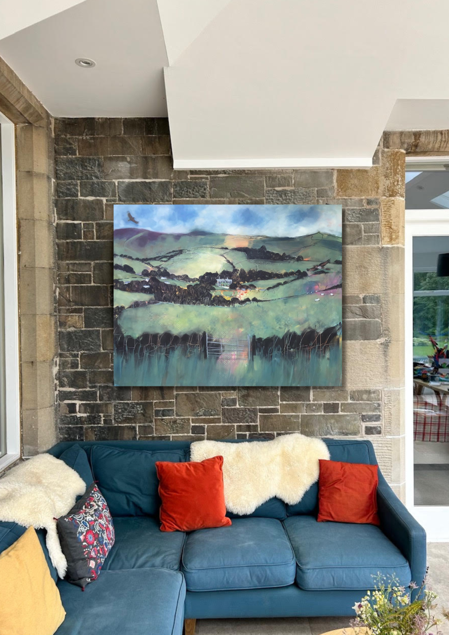 Living room with a blue sofa decorated with red and yellow cushions and white sheepskin throws, a painting of a countryside landscape hung on a brick wall, and a sliding glass door showing greenery outside.