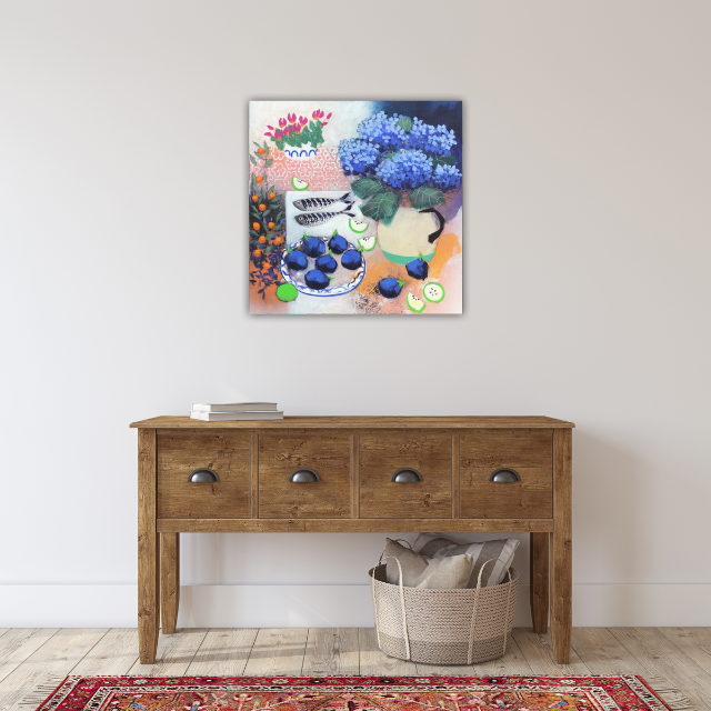 Colorful still life painting featuring flowers, fish, and fruit hanging above a wooden console table with a woven basket and a small stack of books, on a white wall in a room with a red patterned rug.