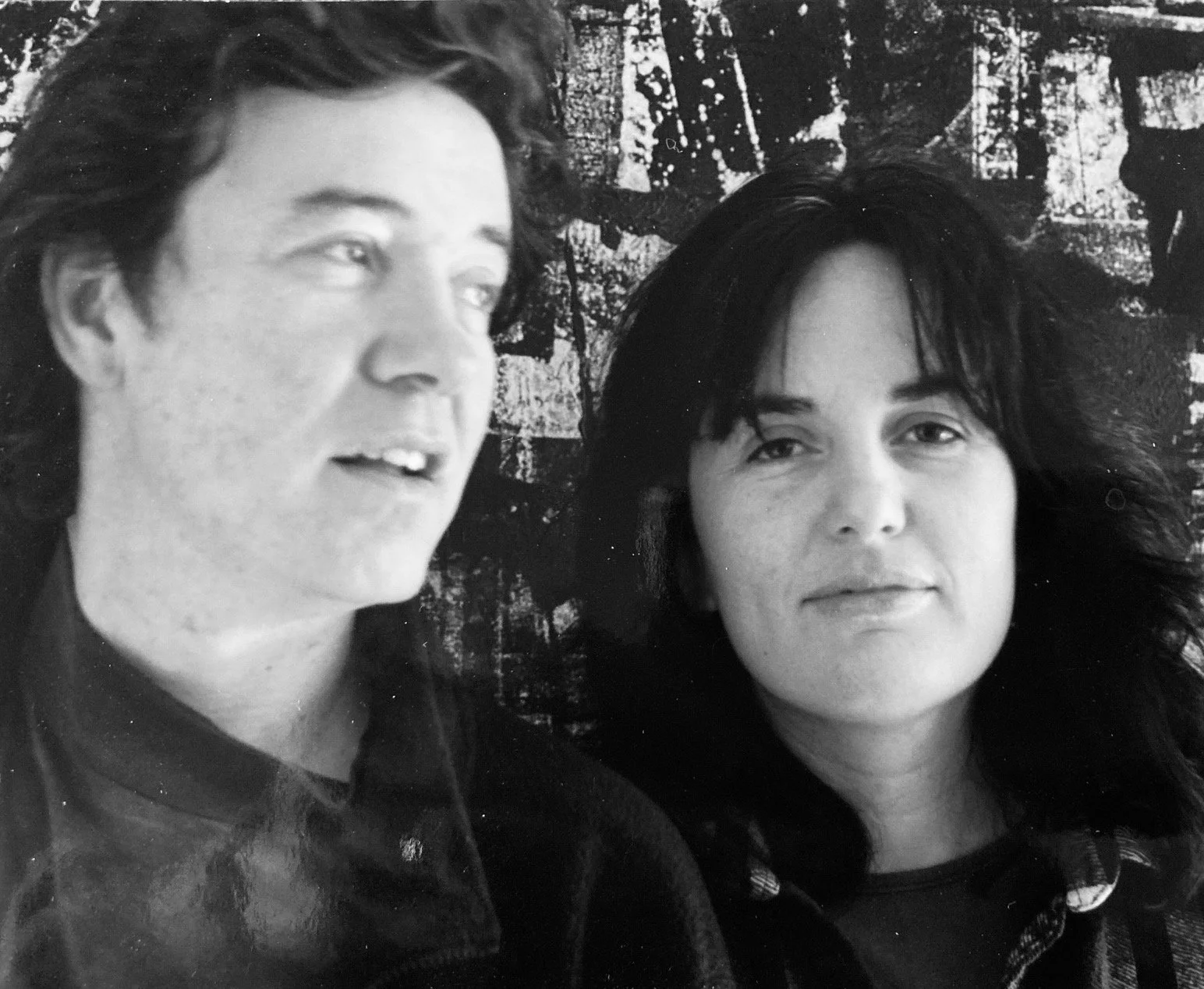 Black and white photograph of a man and woman standing close together, with a textured wall behind them. The man has wavy hair and is looking slightly upward, while the woman has straight, dark hair and is looking directly at the camera.