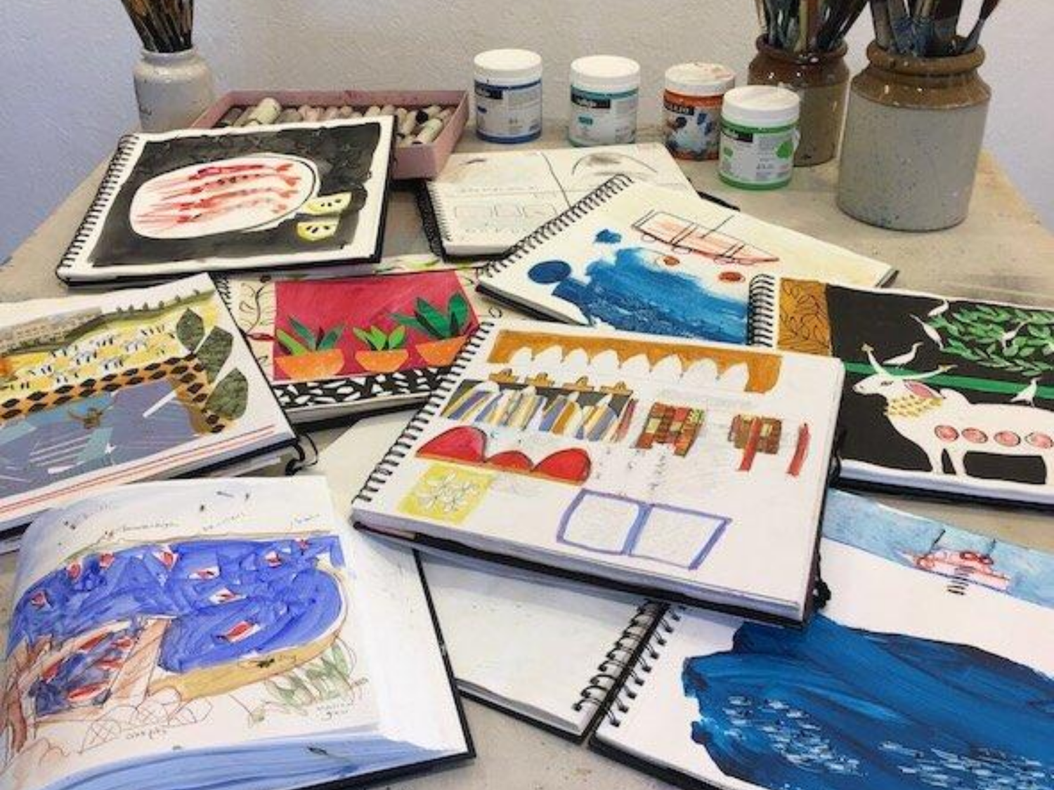 Table covered with open sketchbooks featuring colorful watercolor paintings and illustrations, with paint jars and brushes in the background.