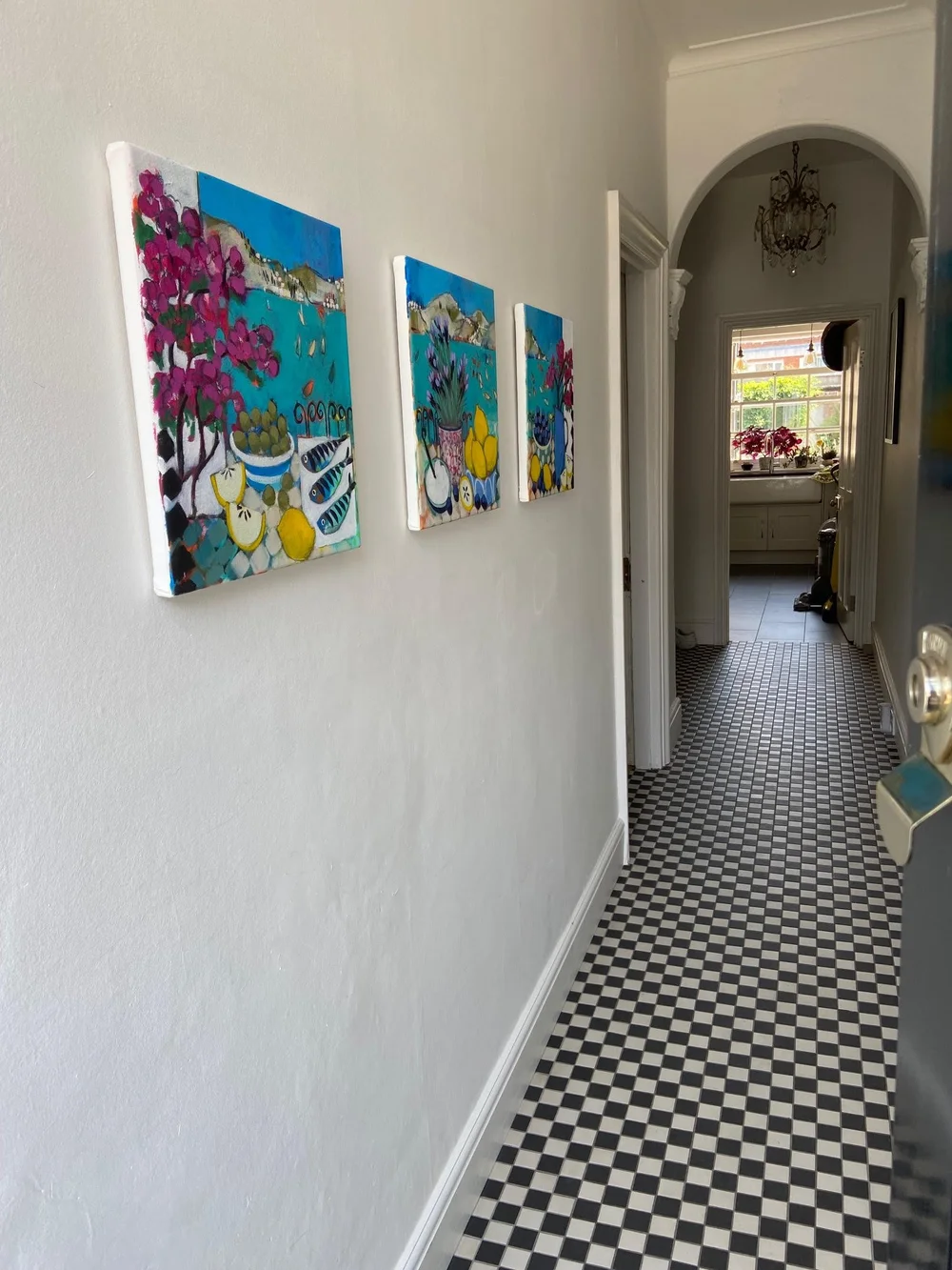 Hallway with three colorful landscape paintings on the left wall, black and white checkered floor, leading to a room with sunlight, pink flowers on windowsill, and chandelier