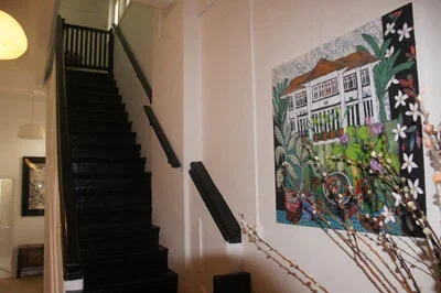 Interior view of a staircase with black steps and railing, and a colorful painting of a house and garden hanging on the wall.