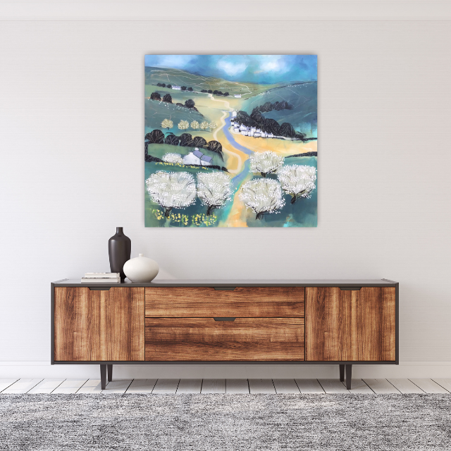 Colorful landscape painting of rolling green hills, a winding river, and blossoming white trees, hung above a mid-century modern wooden sideboard with decorative vases and books.