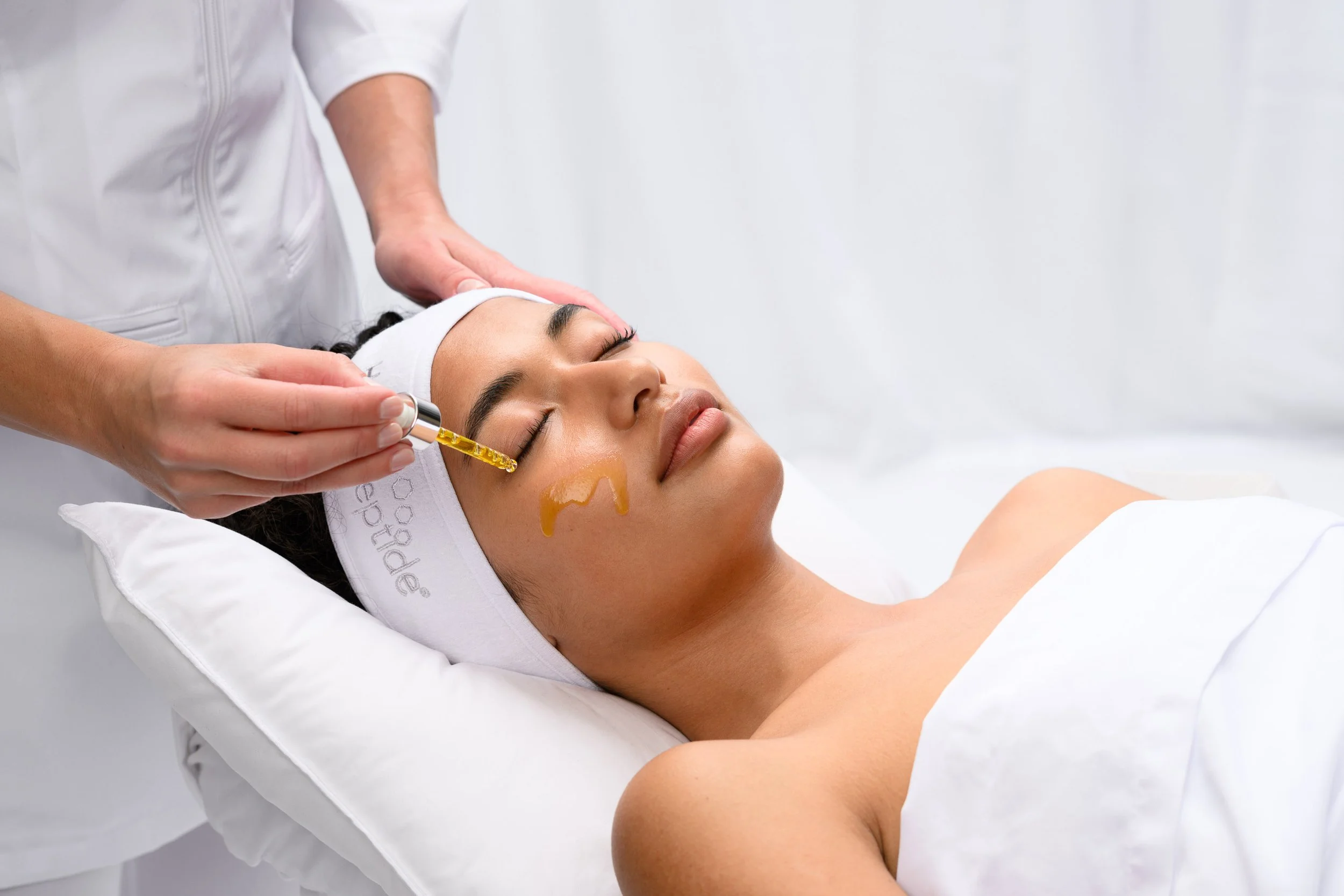 A woman receiving a facial treatment with a skincare serum or oil being applied using a dropper, while lying on a treatment bed with a white headband and covered with a white towel.