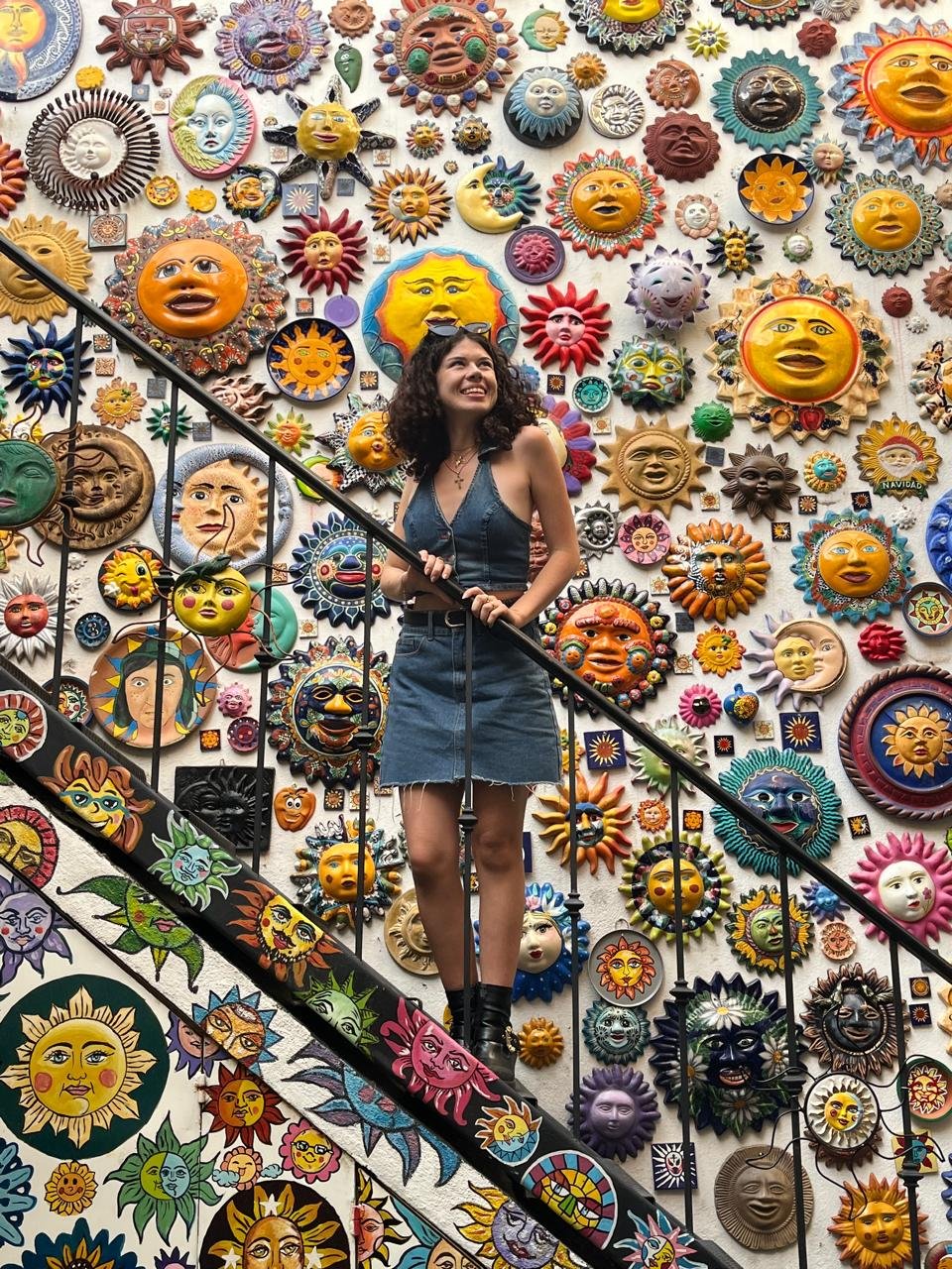 Paola Rousseau Charny, an interior designer is standing on a staircase in front of a colorful wall decorated with various sun and moon face sculptures. Interior design to create functional and colorful spaces.