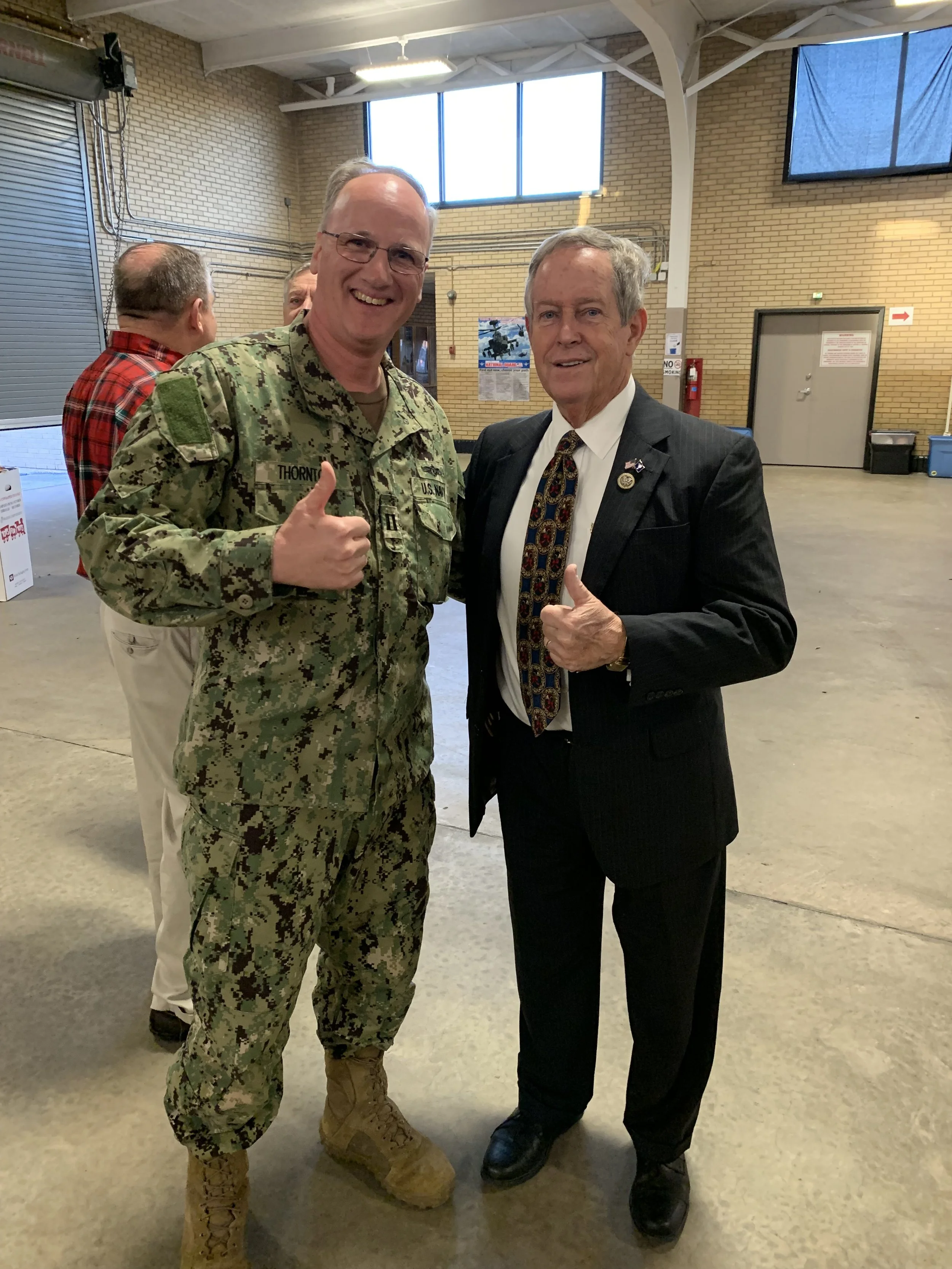 Two men smiling and giving thumbs up, one dressed in military uniform and the other in a suit, standing inside a large indoor space with brick walls and high ceilings.