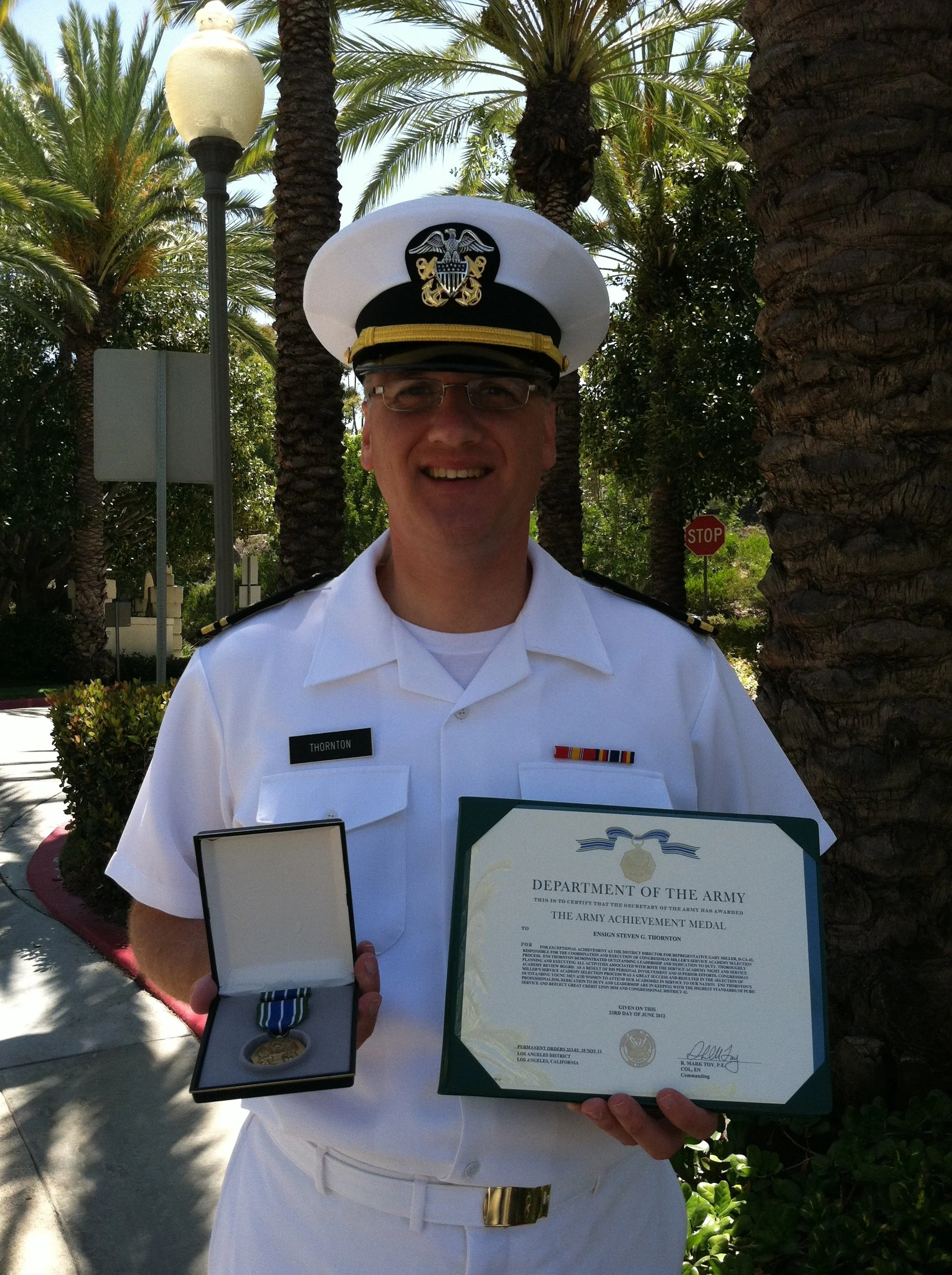 A man in a white military uniform with a cap, holding a medal in a box and a certificate of achievement from the Department of the Army, standing outdoors among palm trees.