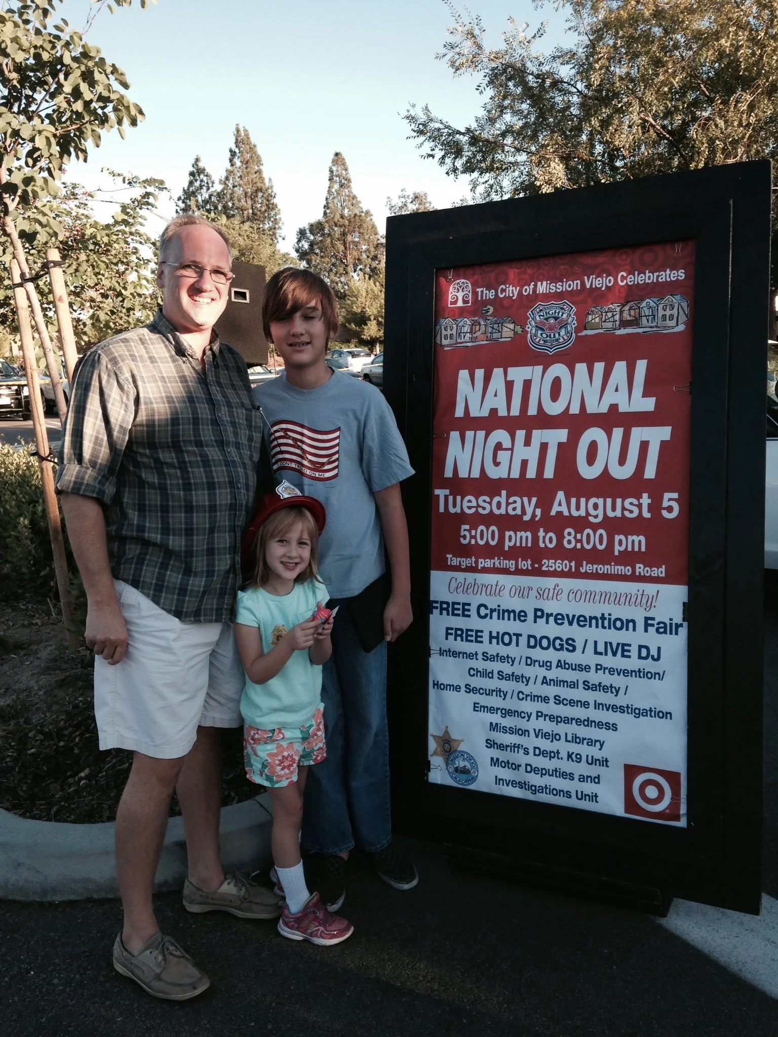 Three people, an adult male and two children, standing next to a sign board for a National Night Out event in Mission Viejo. The sign displays details about the event on Jeronimo Road.