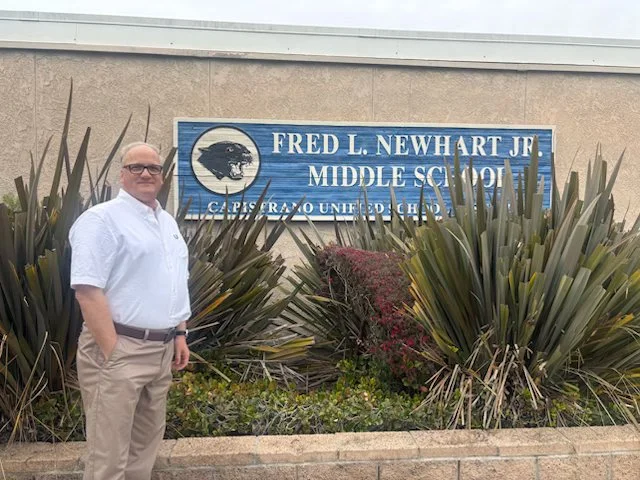 Man standing in front of a sign that reads 'Fred L. Newhart Jr. Middle School, Capistrano Unified School District,' with desert plants around the sign.