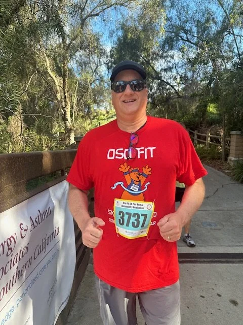 Smiling man in red ASOFT t-shirt, dark sunglasses, black hat, and race bib number 3737, standing outdoors on a trail or park path.