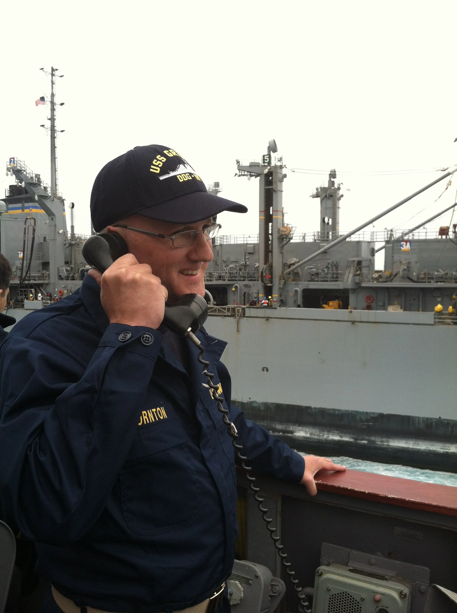 A sailor talking on a ship's communication device on a gray naval vessel, wearing a navy uniform and cap with yellow lettering.