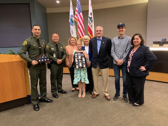 Group of seven people standing in front of flags in a room, including police officers, children, and adults, with a desk on the left.