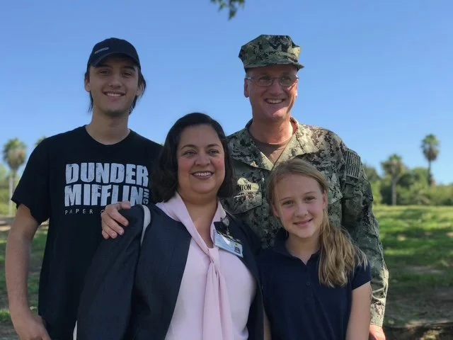 Four people, including a soldier in camouflage uniform and glasses, standing outdoors with a blue sky and palm trees in the background, smiling for a photo with two young girls and a woman.