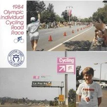 Poster for the 1984 Olympic Individual Cycling Road Race featuring a cyclist, traffic cones, and a child smiling at the camera, with signs indicating La Paz and cycling directions.