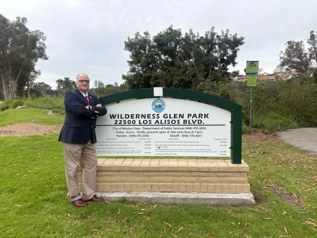 Man in a suit standing next to a sign for Wilderness Glen Park on Los Alisos Boulevard.