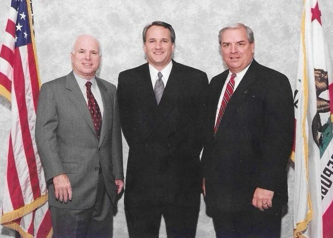 Three men in suits standing in front of American and California flags.