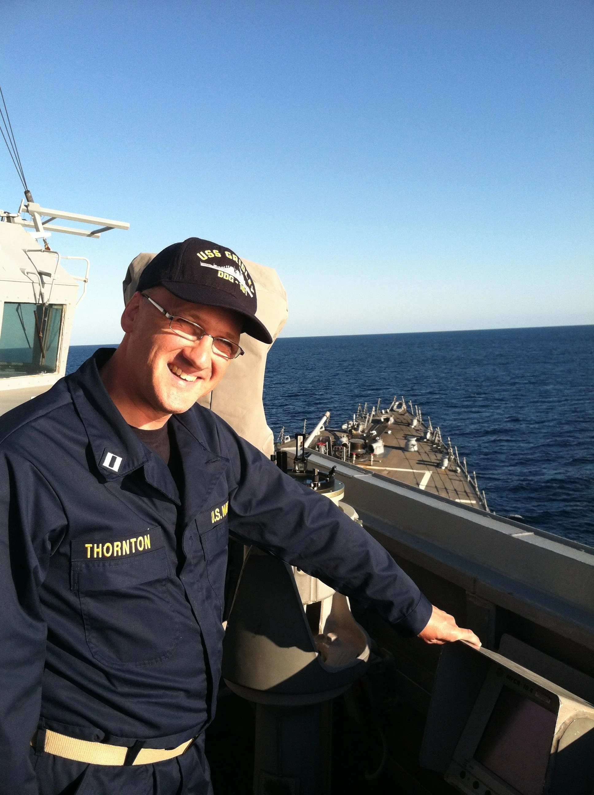 A man in a U.S. Navy uniform on a ship's bridge, smiling and looking at the camera with the ocean in the background.