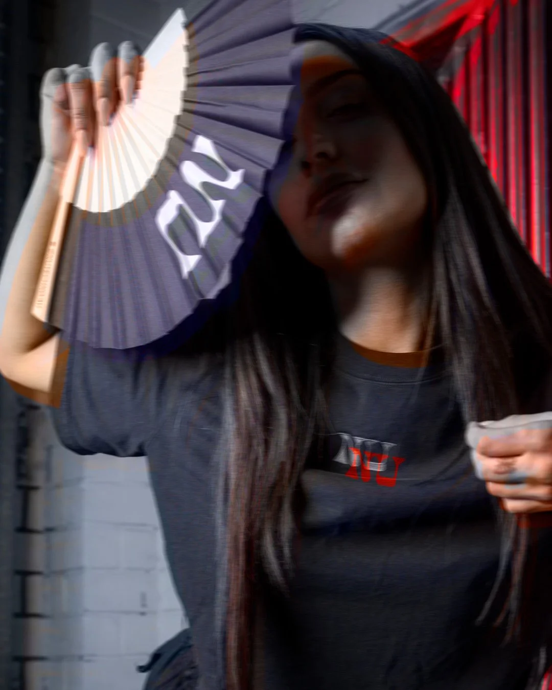 A woman with long dark hair holds a blue and white fan with the word "LOVE" on it, partially obscuring her face, with red curtains in the background.