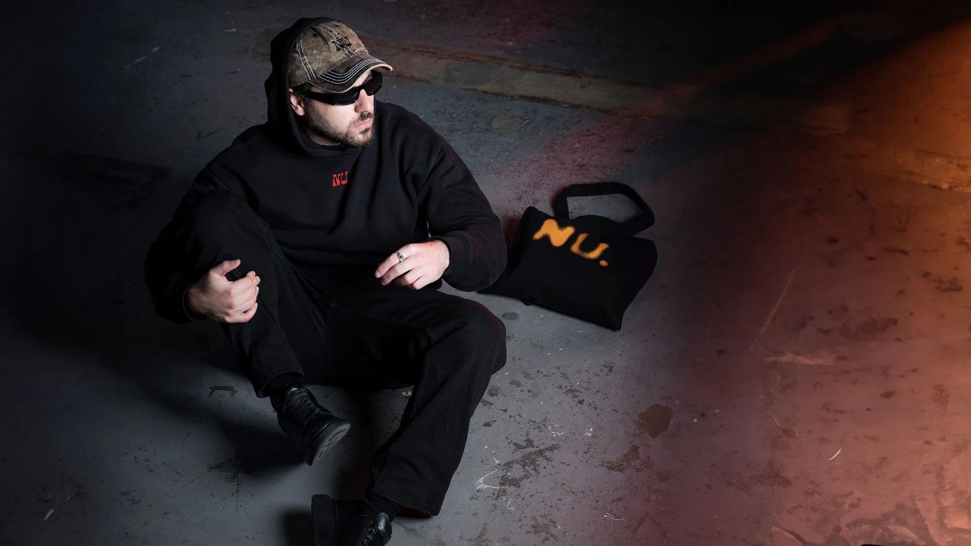 A man dressed in black, wearing sunglasses and a camouflage cap, is sitting on the floor in a dark, industrial-like space. He is holding his knee with one hand. Next to him on the floor is a black bag with the letters "NU" in yellow.