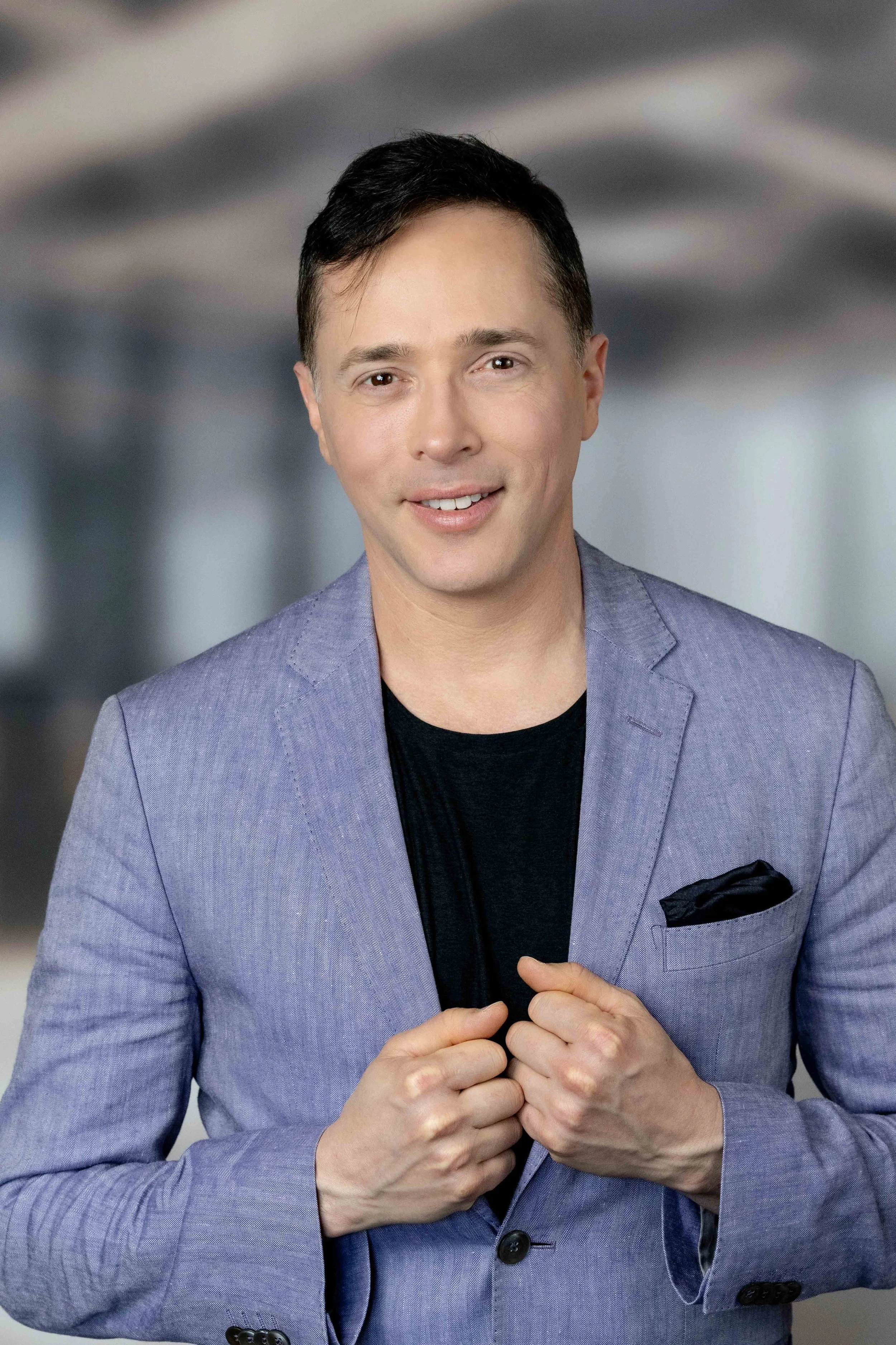 Yuval David, in a gray blazer adjusts his lapel, smiling, in a professional portrait setting.