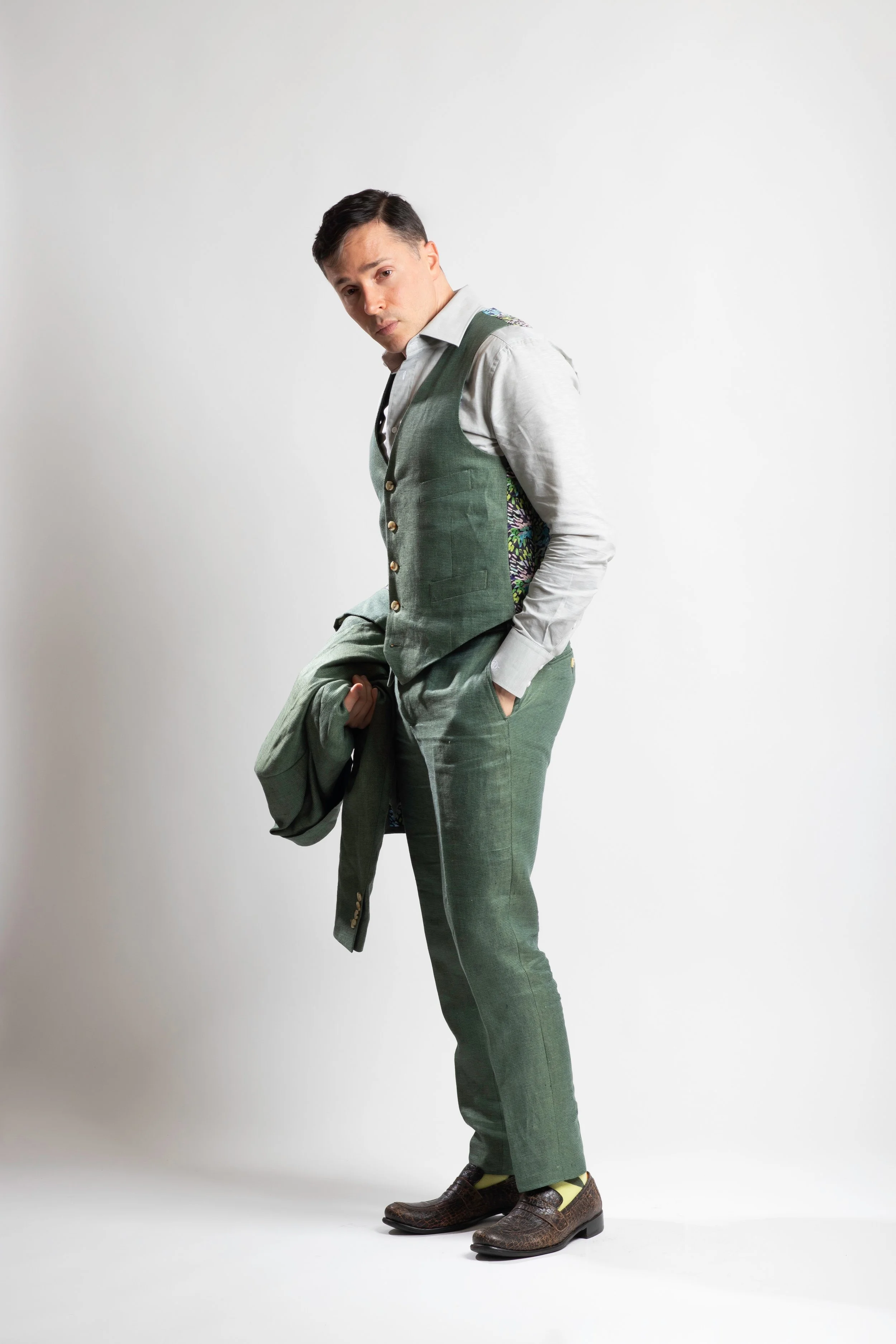 Yuval David, in a light-colored shirt, green vest, and green pants stands against a white background, holding a matching green jacket in his hand and looking at the camera.