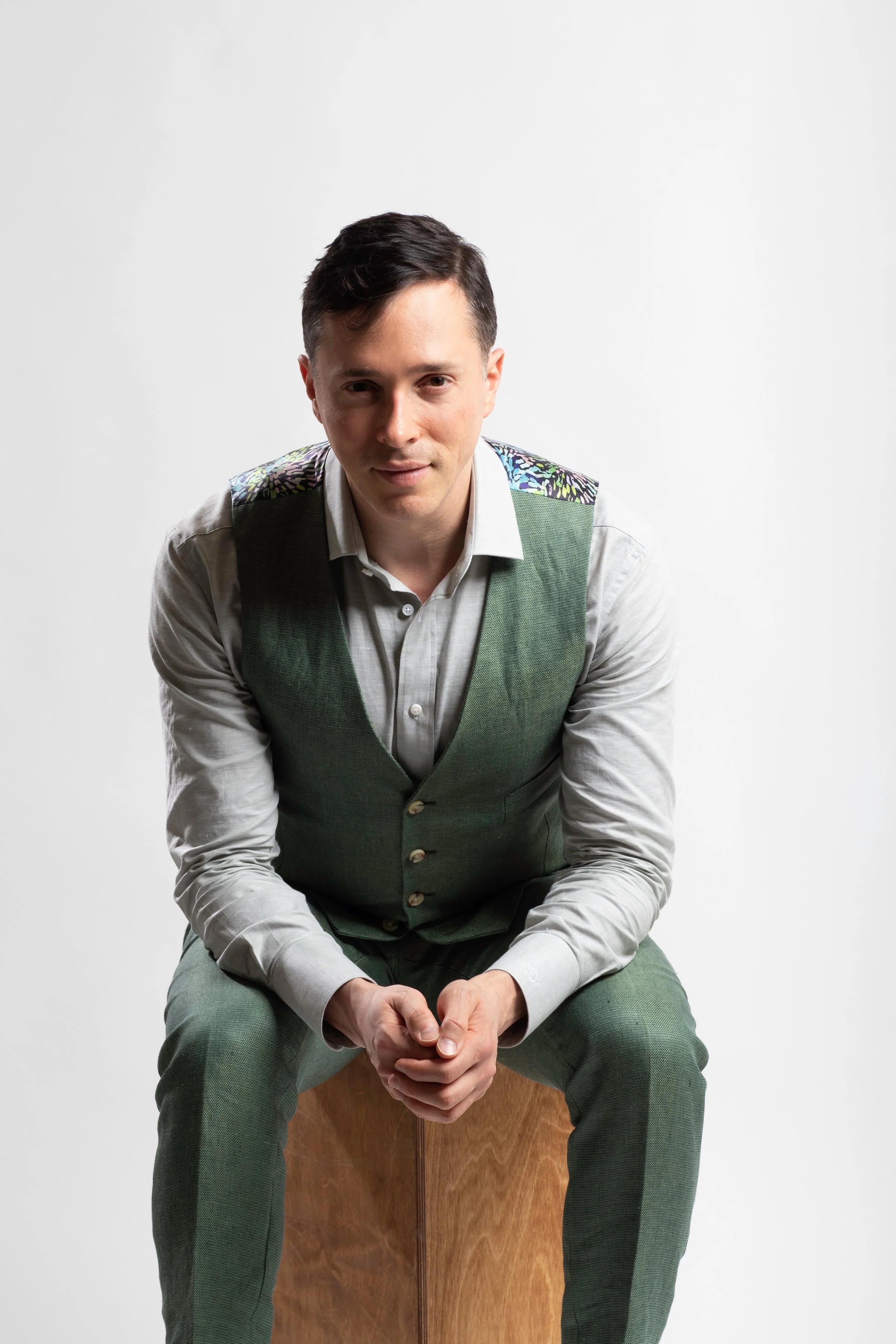 Yuval David, wearing a light gray shirt and a green vest with decorative shoulder patches, sitting on a wooden cube against a white background.