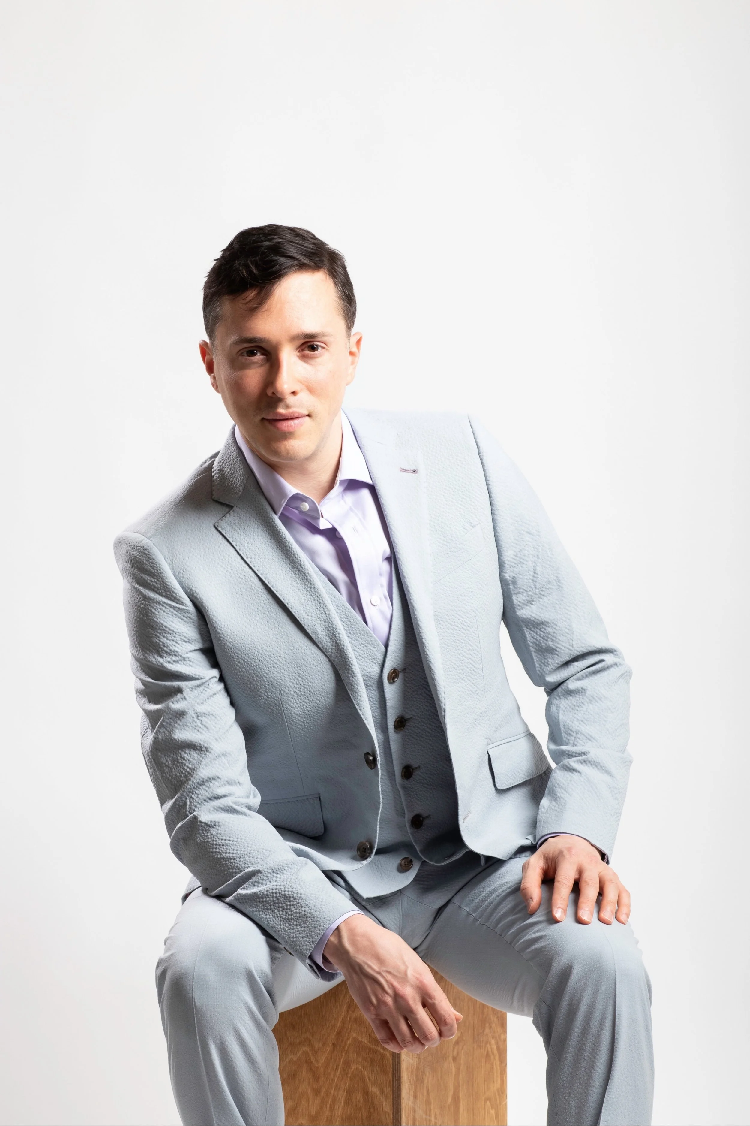 Yuval David, dressed in a light gray suit, sitting on a wooden box against a plain white background.