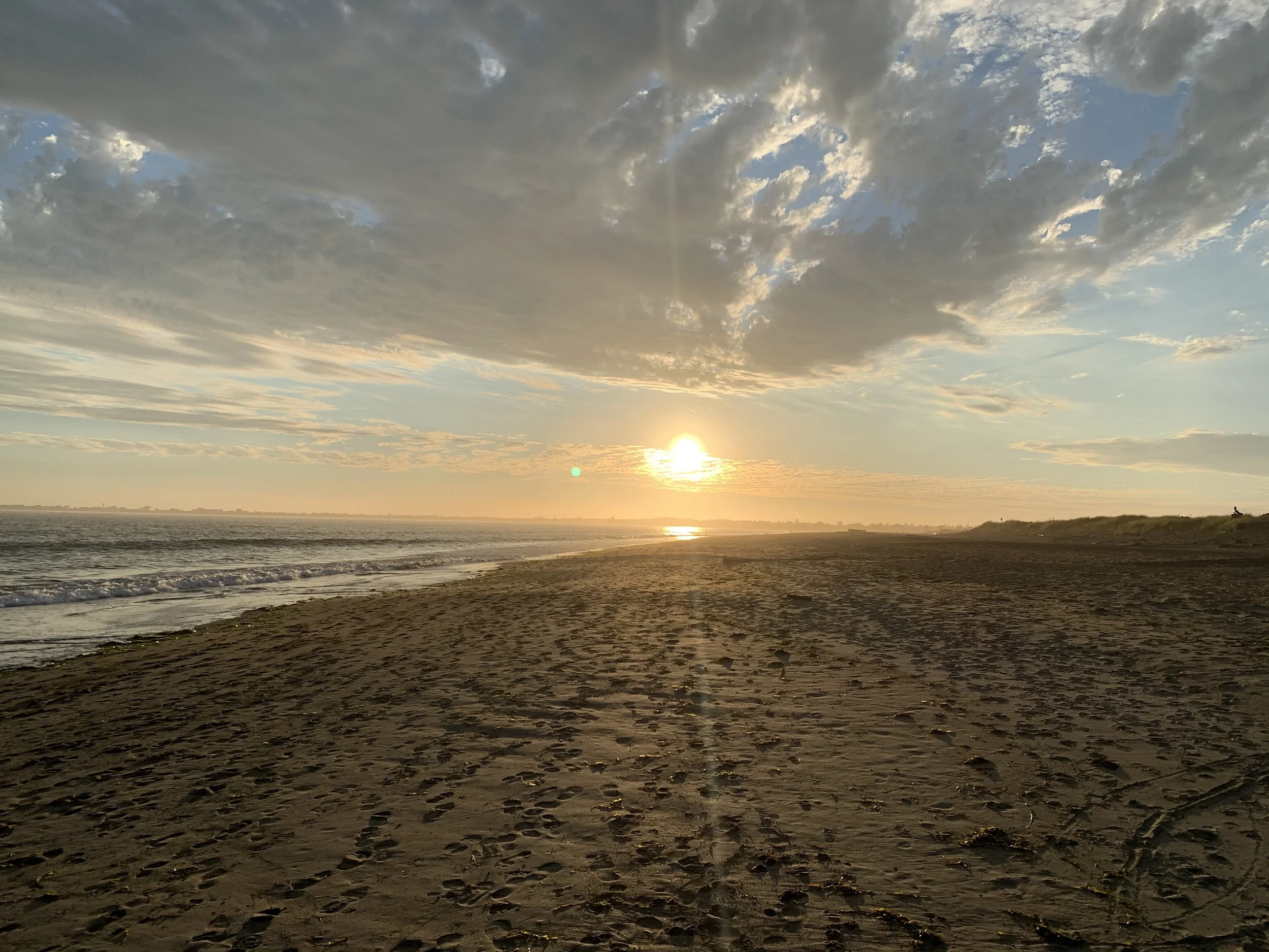 A beach at sunset with footprints in the sand, calm waves, and partly cloudy sky with the sun near the horizon.