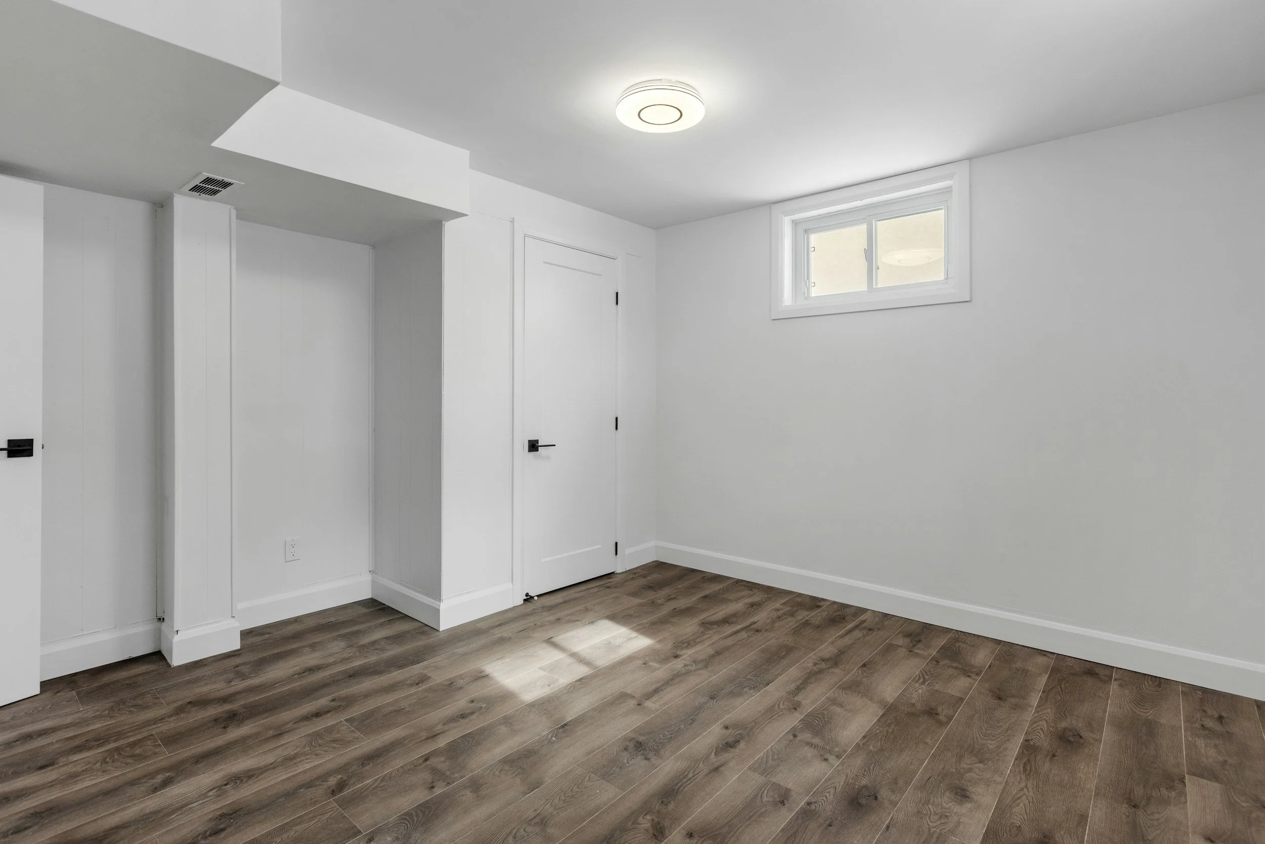 Empty room with white walls, a small rectangular window, a ceiling light, and wood-look flooring.