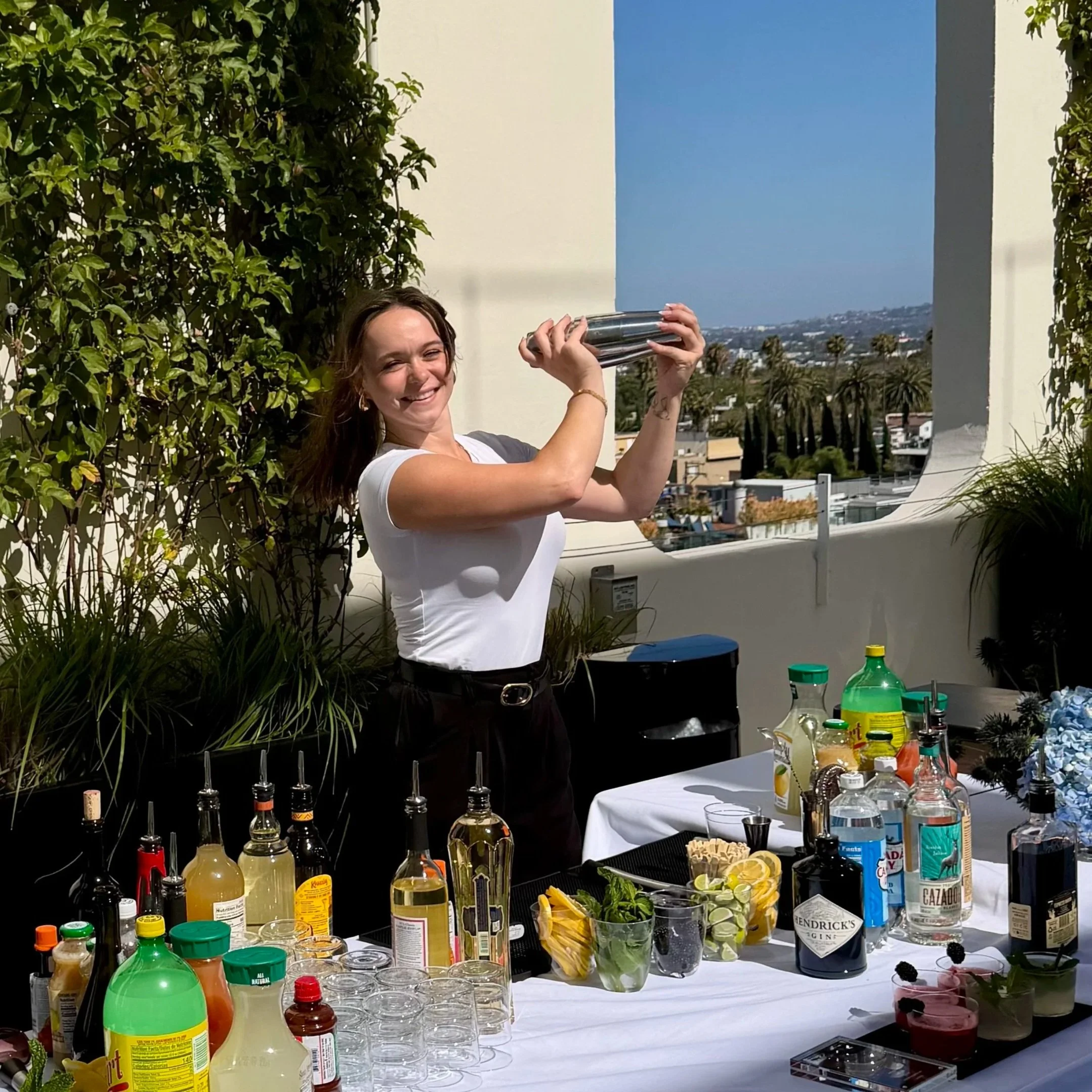 A woman smiling and holding a cocktail shaker standing behind a table with various bottles of alcohol, mixers, and garnishes, at an outdoor event with palm trees and a city view in the background.