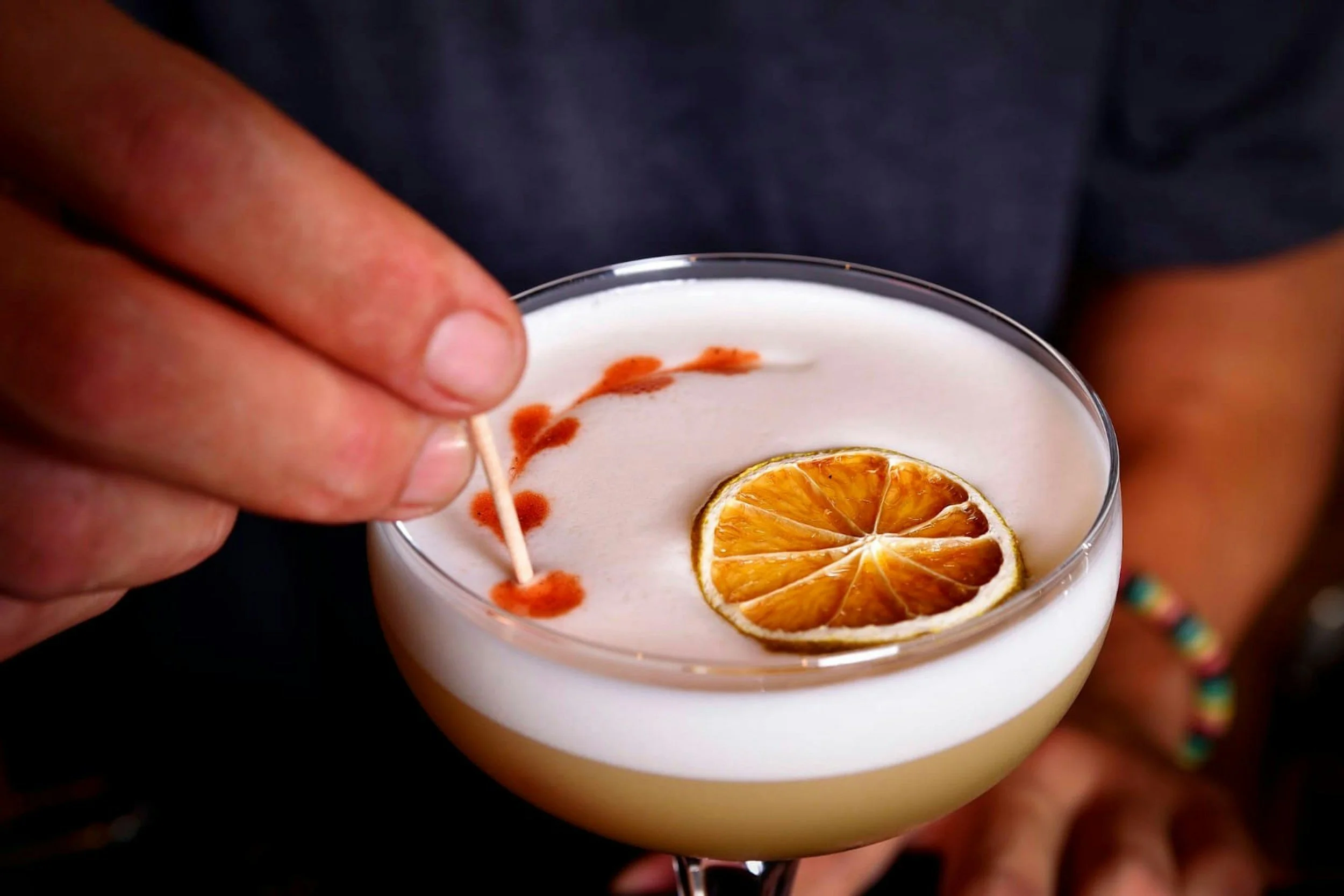 Close-up of a cocktail in a glass with a dried orange slice garnish, a cinnamon stick, and red spice powder decoration on the surface.