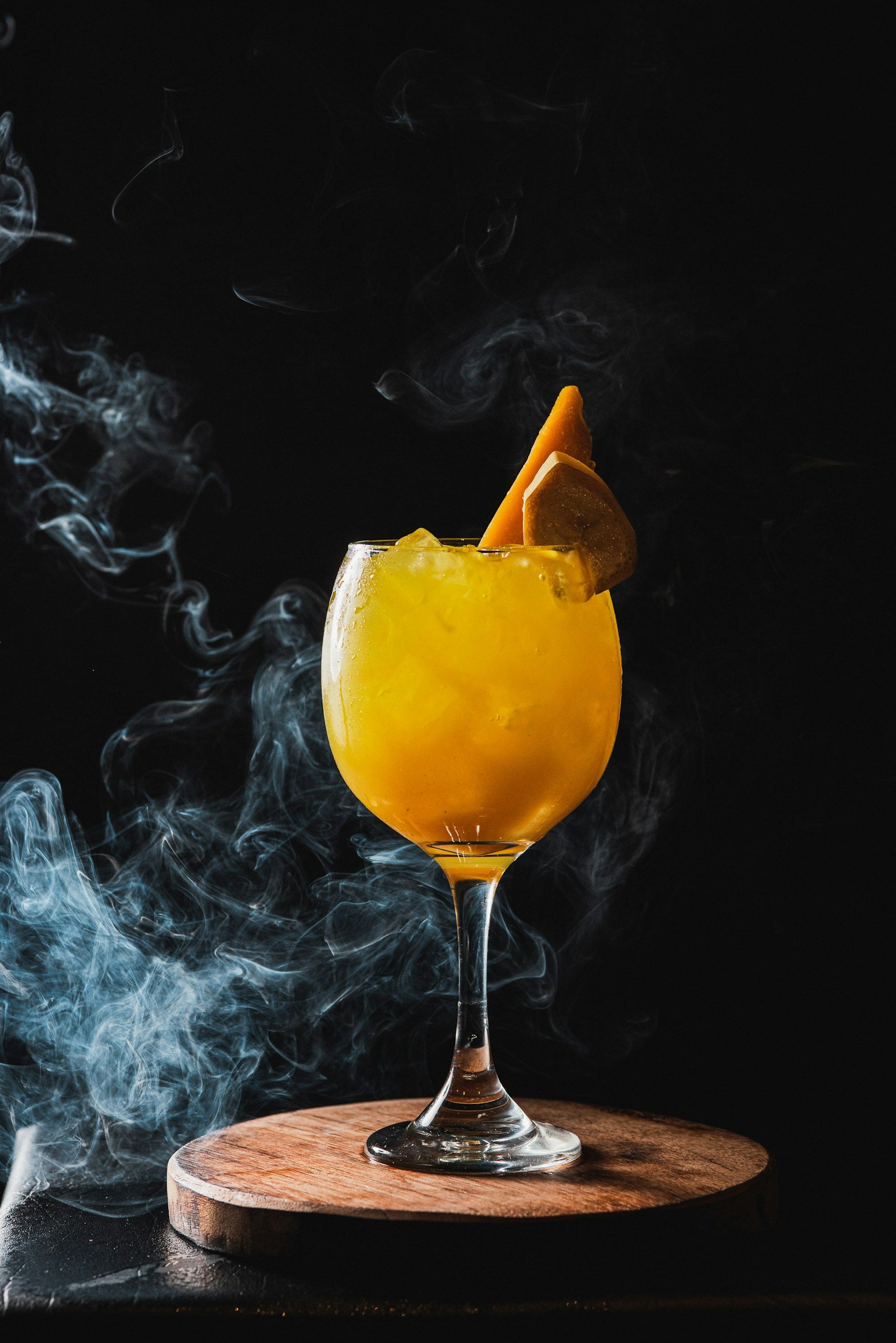 A yellow cocktail in a glass garnished with a slice of orange and a wedge of lemon, with wisps of smoke swirling around on a dark background.