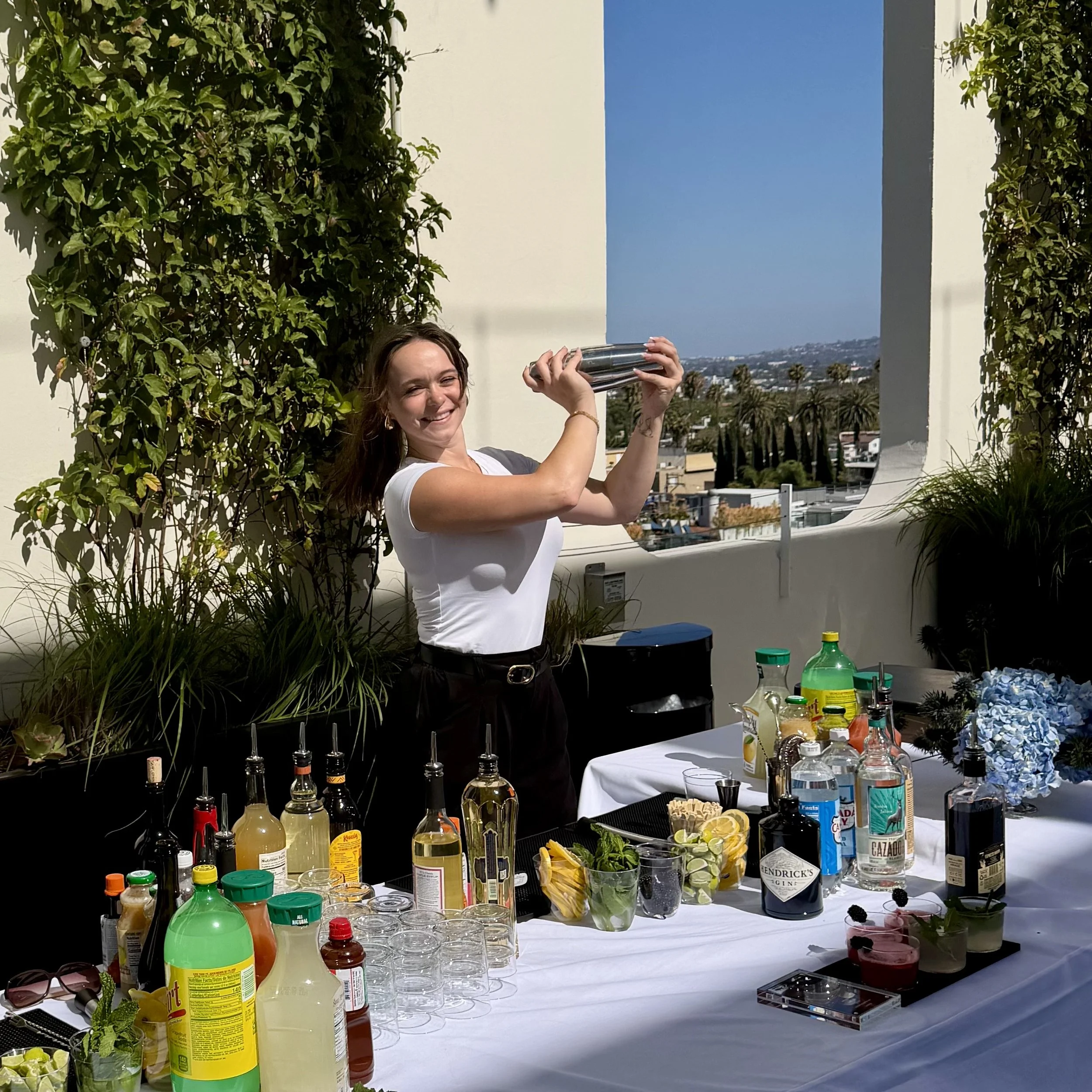 A woman with brown hair is smiling and holding a cocktail shaker, standing behind a table with various bottles, glasses, and ingredients for making drinks. The setting appears to be an outdoor event on a balcony with a cityscape and palm trees in the background.