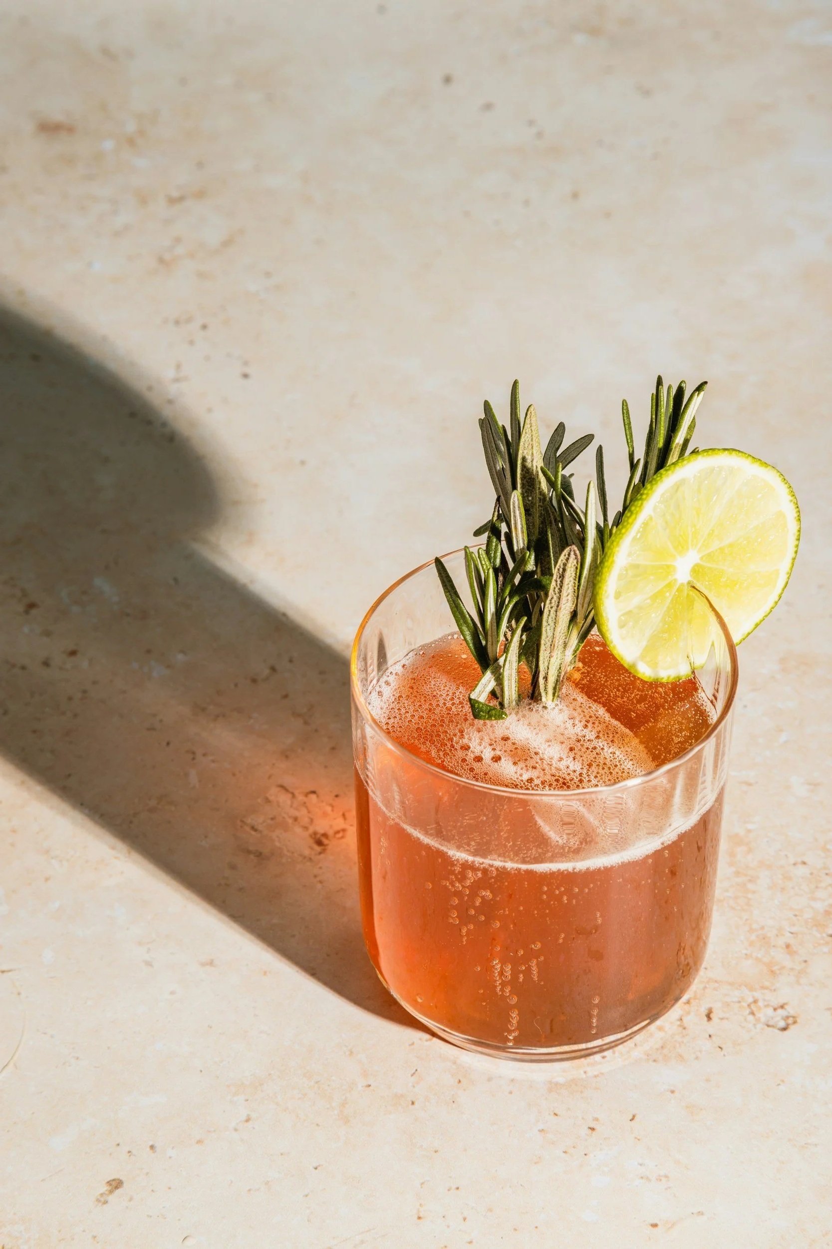 A pinkish-orange cocktail drink in a clear glass, garnished with a lemon slice and sprigs of rosemary.