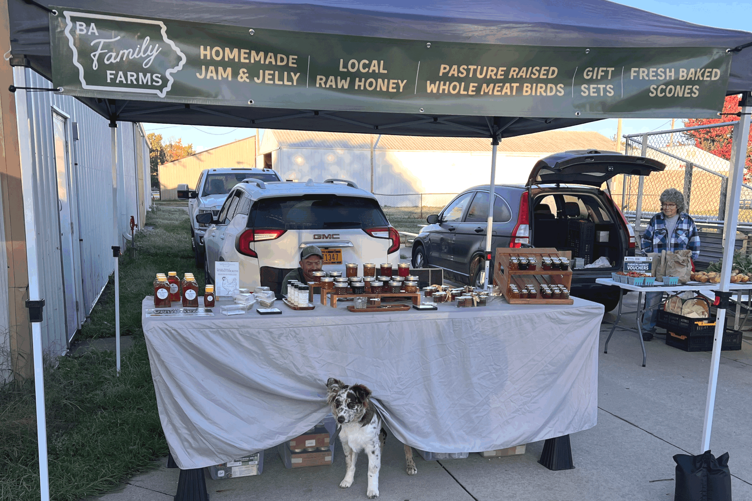 An outdoor market stall with a green canopy displaying farm products such as homemade jam and jelly, local raw honey, pasture-raised whole meat birds, gift sets, and fresh baked scones. Two people are behind the table, one smiling and the other working. A dog stands in front of the table. Several cars are parked behind the stall.